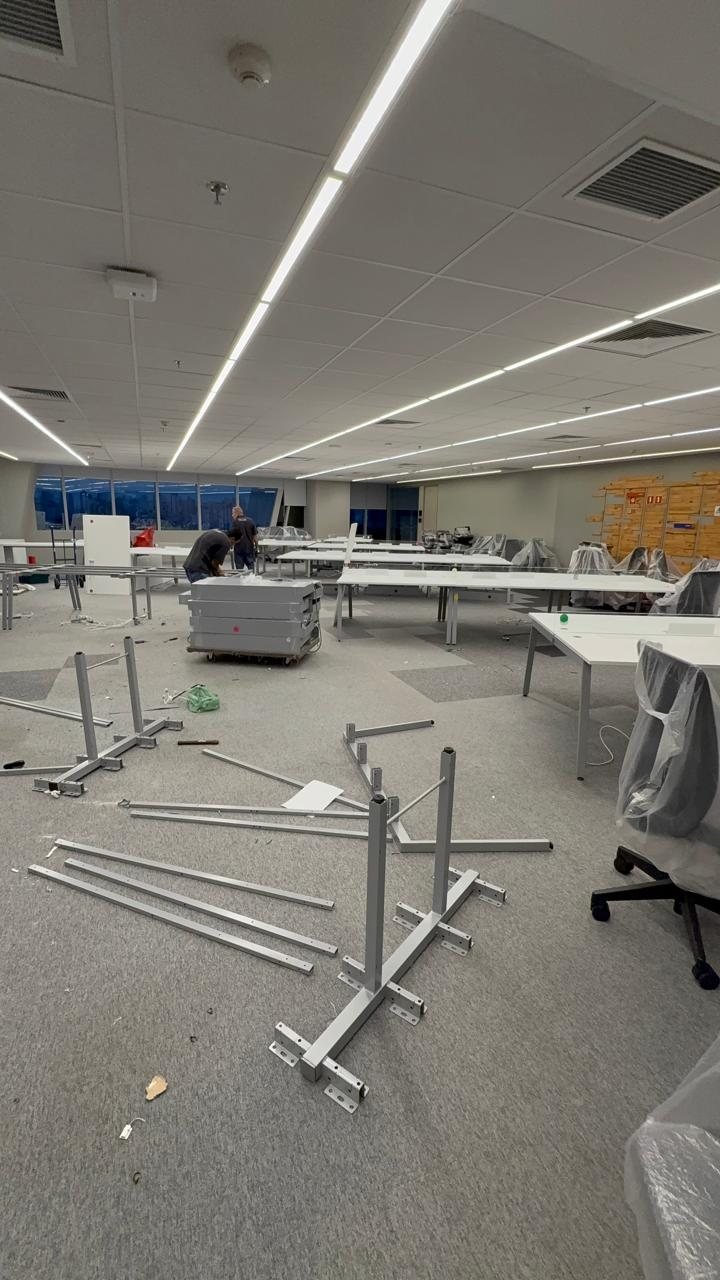 Montagem em andamento: estruturas de mesa desmontadas no piso, tampos protegidos com plástico e equipe trabalhando ao fundo.